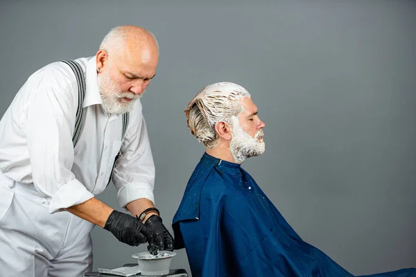 Cabeleireiro sênior aplicando tintura ao cabelo do homem. Processo de um tipo ter o cabelo pintado no cabeleireiro. Conceito de beleza e pessoas. Homem estilista com tintura de cabelo e cabelo escova para colorir no salão. — Fotografia de Stock