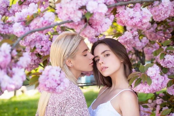 Unga flickor och vårblommor. Porträtt av två vackra unga kvinnor som kopplar av i sakura blommor. Lesbiska par kysser varandra. Känslig beröring och kyss. Flickvänner par. — Stockfoto