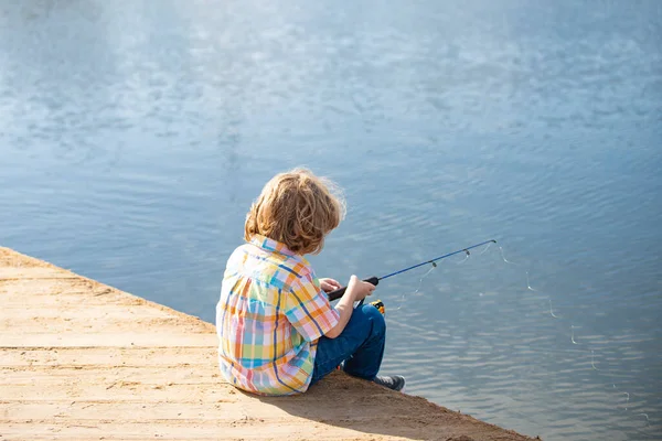 Kinderangeln. Jungfischer. Junge mit Spinner am Fluss. Porträt eines aufgeregten Jungen beim Angeln. Junge am Steg mit Rute. Fischereikonzept. — Stockfoto