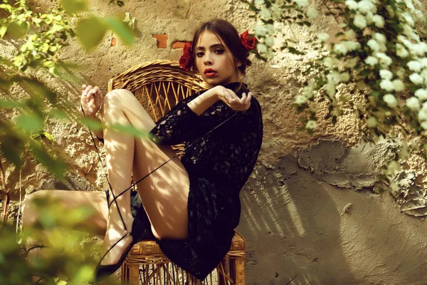 Дівчина зав'язує шнурки взуття на плетеному кріслі в сонячний день. Стильна модель в модному чорному платті, макіяж червоних губ і троянди в брюнетці волосся. Мода. Весна, літо. Квіти, цвітіння . — стокове фото