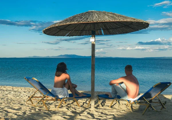 Pareja joven sentada en una arena y mirando a un mar bajo el paraguas. Verano relajarse en la playa. — Foto de Stock