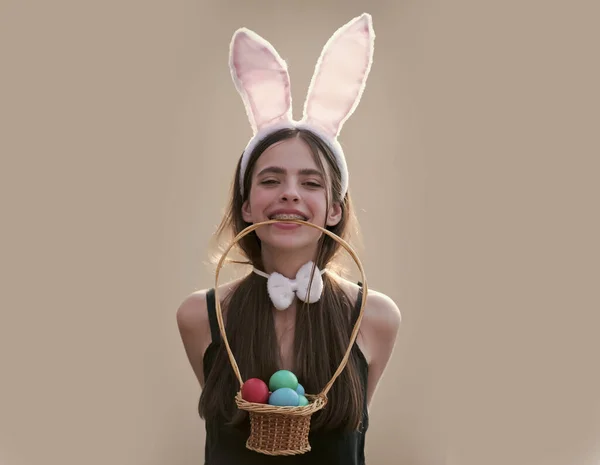 Wielkanocna kobieta z króliczymi uszami i kokardą na beżowym tle. Dziewczyna trzyma wiklinowy koszyk z jajkami w zębach. Płodność i koncepcja odrodzenia. Wiosenne święto. Wielkanoc tradycja i symbol. — Zdjęcie stockowe