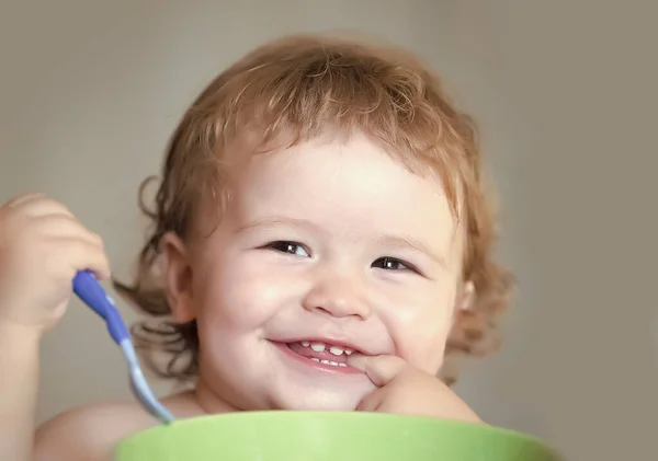 Baby eating. Portrait of funny little smiling boy with blonde curly hair and round cheecks eating from green plate holding spoon closeup. — Stock Photo, Image