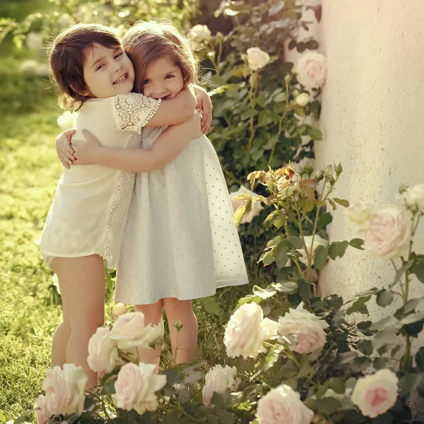 As crianças que sorriem para flores de rosa florescentes. Duas meninas abraçando no jardim de verão no dia ensolarado. Amigos e amizade. Feliz conceito de infância. Germinação e florescimento. — Fotografia de Stock