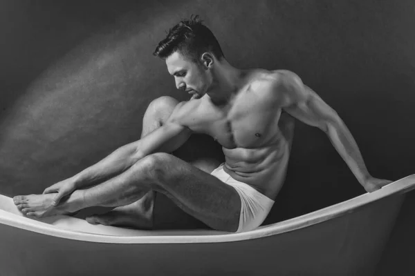 Junger Mann in weißen Höschen mit sexy nacktem muskulösen Oberkörper, der auf einer Badewanne sitzt. Mans Unterhosen und Höschen. — Stockfoto