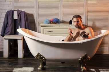 Yüzünde köpük olan sakallı genç bir adam jiletle küvette tıraş oluyor. Sağlık güzelliği ve cilt bakımı konsepti. Banyodaki adam..