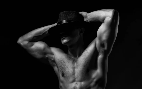 Muž ve stínu. Mladý muž s svalnatým tělem v klobouku s holou hrudí a trupem pózující ve studiu na černém pozadí. — Stock fotografie
