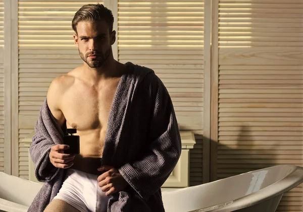 Bărbat cu trunchi muscular în lenjerie intimă și halat de baie în baie. Tipul cu sticla de parfum in baie . — Fotografie, imagine de stoc