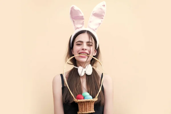 Chica de Pascua sosteniendo cesta con huevos en la boca. Mujer con orejas de conejito rosado sobre fondo beige. Concepto de fertilidad y renacimiento. Tradición y símbolo de Pascua. —  Fotos de Stock