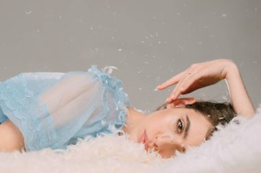 Sakin yüzlü kız tüy ve tüylerle kaplı olarak yatağa uzanıyordu. Yumuşak ve nazik bir kavram. Tüyler uçuşurken mavi gecelikli kadın yatakta. Şefkatli gençlik kavramı.