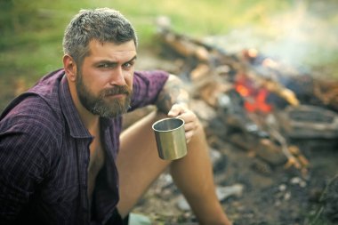 Elinde kupa olan bir gezgin doğa üzerindeki şenlik ateşinde rahatlar. Hippi yürüyüşçü kamp ateşinde çay içer. Kamp ve yürüyüş, yaşam tarzı. Seyahat kavramı.