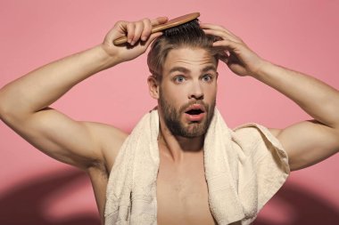 Şaşırmış suratlı, göğsünde banyo havlusu olan bir adam. Pembe arka planda saç fırçası olan bir adam. Saç bakımı, sağlık. Sağlık saçı ve hijyen. Sabah rutini konsepti.