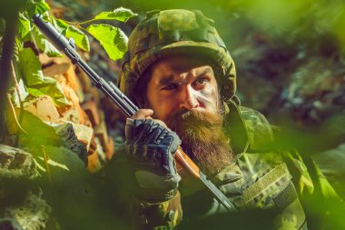 Askeri miğferli, üzgün yüzlü ve silahlı bir asker. Tuğlalara dayalı bir kamuflaj. Ormanda..