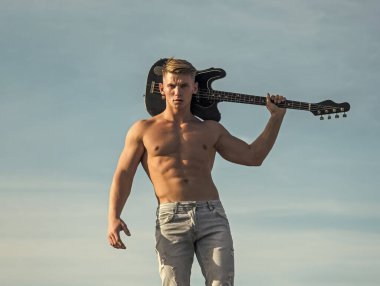 Altılı paketli bir atlet gitarist, açık havada kot pantolon giymiş. Müzik, performans, eğlence. Gövdesi kaslı bir adam elektro gitarı havada tutuyor..