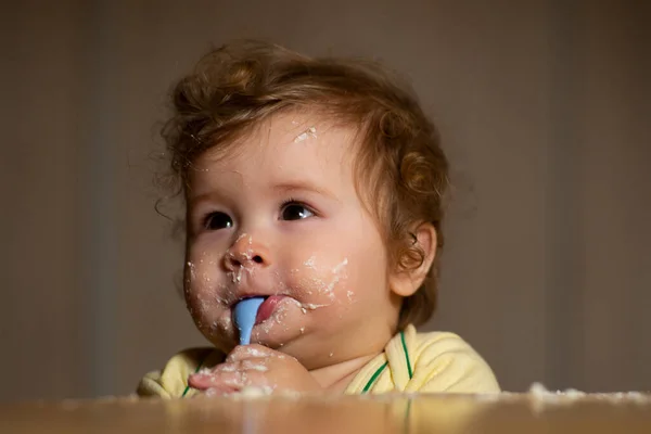 Kids baby eating with dirty face. Cheerful smiling child child eats itself with a spoon Baby eating with dirty face. Smiling little child eating food on kitchen. Kid hungry so parent feed him.