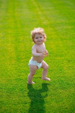 İlk adım. Bebek bez pantolonuyla yeşil çimlerde çıplak ayakla duruyor. Güzel, mutlu küçük çocuk yaz parkında yeşil bir çayırda yürüyor..