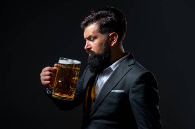 Klasik takım elbiseli adam bira içiyor. İş kıyafetli sakallı adam mutlu ve memnun görünüyor. Siyah arka planda yüksek bardak bira taşıyan bir adamın portresi..