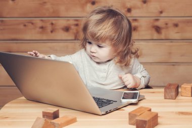 Bilgisayarda ve cep telefonuyla oynayan sevimli, komik bir çocuk. Oyuncak bina bloklarının yanında, ahşap arka planda..