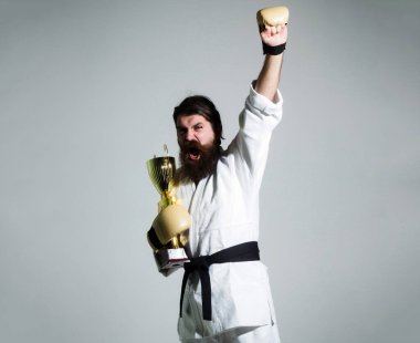 Beyaz kimonolu komik karateci beyaz hippi siyah kemerli ve boks eldivenli neşeli yüzlü altın şampiyon kupasını gri arka planda tutuyor..