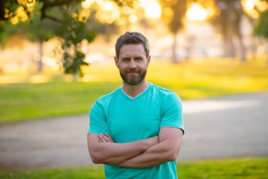 Parkta kollarını kavuşturmuş tişörtlü yakışıklı bir genç..