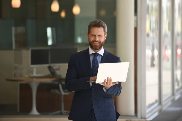 Επιχειρηματίας κρατώντας το laptop του στέκεται στο κοστούμι στο φόντο του γραφείου. — Φωτογραφία Αρχείου