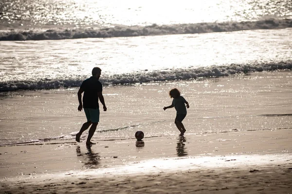 父と息子は夏休みにビーチでサッカーやサッカーをします。父と子供は屋外で楽しんでいます。夏の日に遊んでいる子供の男の子とパパ,日没のシルエット. — ストック写真