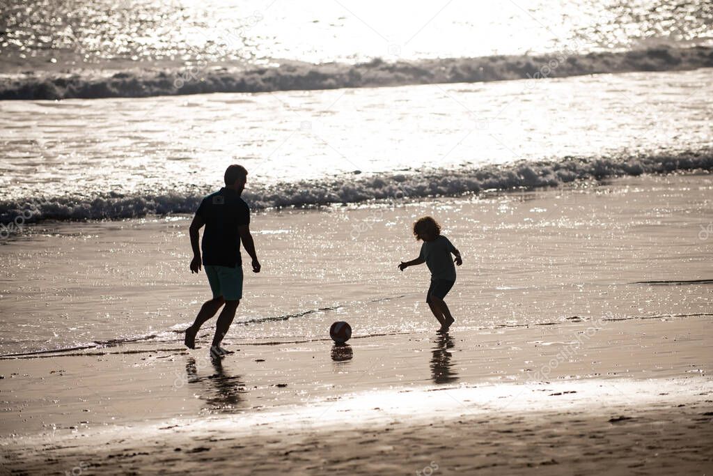 Father and son play soccer or football on the beach on summer family holidays. Dad and child having fun outdoors. Daddy with kid boy playing on a summer day, silhouette on sunset.