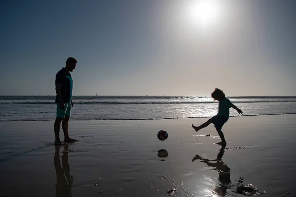 父と息子はビーチでサッカーやサッカーをします。夏の日に遊んでいる子供の男の子とパパ. — ストック写真