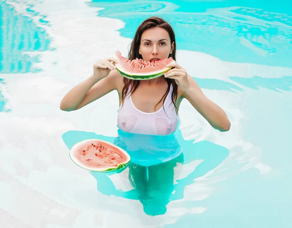 Sexy vrouw in het zwembad met blauw water in natte witte vest met blote borst eten rode watermeloen zonnige zomerdag buiten. — Stockfoto