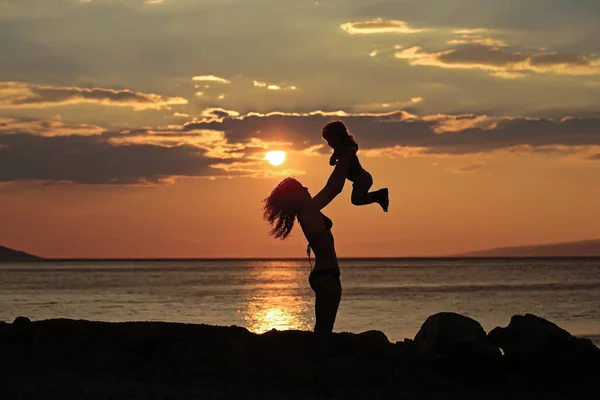 母と赤ちゃんのシルエットは、夜や夕暮れ時に海や海の水の近くのビーチで再生されます自然の背景に夏の日. — ストック写真