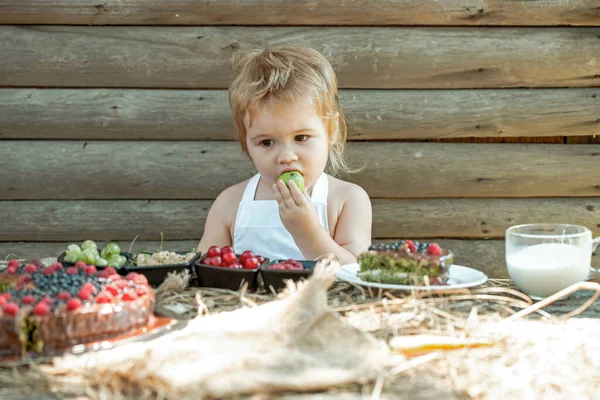 Roztomilé dítě jí zelené jablko u stolu s bobulemi malina angrešt cherry rybíz ovocný dort a šálek mléka venku na dřevěném pozadí. — Stock fotografie