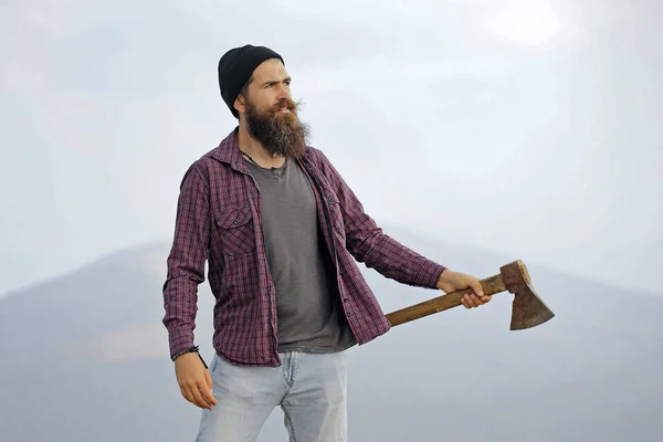 Homme avec hache sur la montagne. Beau hipster homme en chemise à carreaux avec longue barbe tenant hache ou hache sur le sommet de la montagne sur fond naturel avec ciel. — Photo