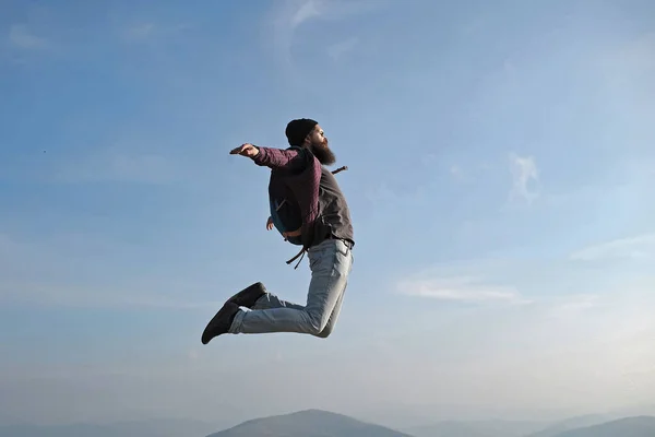 Man hipster jumps with backpack. Carefree happiness freedom concept. Hipster man with beard and moustache in hat jumps with axe on mountain top on cloudy sky. — Stock Photo, Image