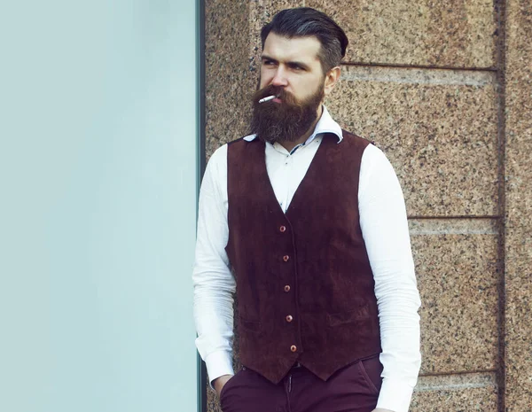 Hombre Hipster fumando cigarrillo. Hombre caucásico con barba en chaleco marrón y pantalones al aire libre en la calle de la ciudad. Estilo de vida urbano. — Foto de Stock