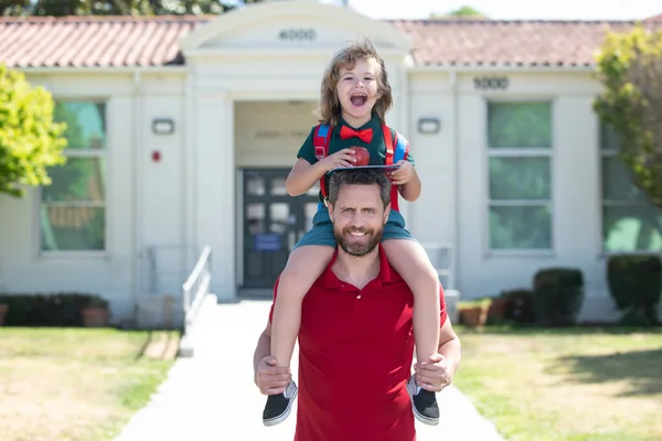 Amerikan baba ve oğlu okul parkında yürüyor. Erkek ve heyecanlı çocuk okulun yanında sırtına biniyor.. — Stok fotoğraf