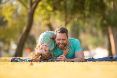 Baba ve oğul doğa parkında çimlerin üzerinde yatıyor. İnsanlar dışarıda eğleniyor. Mutlu tatiller ve dost canlısı aile kavramı.