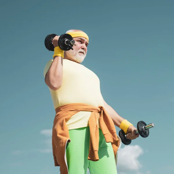 Senior male is enjoying sporty lifestyle. Old mature man exercising with dumbbell. Senior man lifting weights. Grandfather sportsman portrait on blue sky backgrounds.