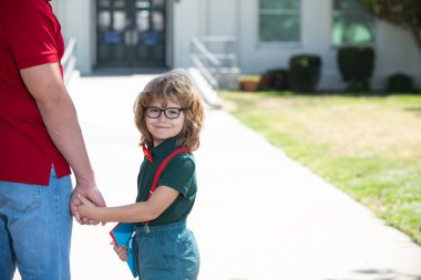 Öğretmenler günü. İlkokulun ebeveynleri ve öğrencileri el ele gider. Tişörtlü öğretmen ve okul parkının yanında sırt çantalı sevimli okul çocuğu..