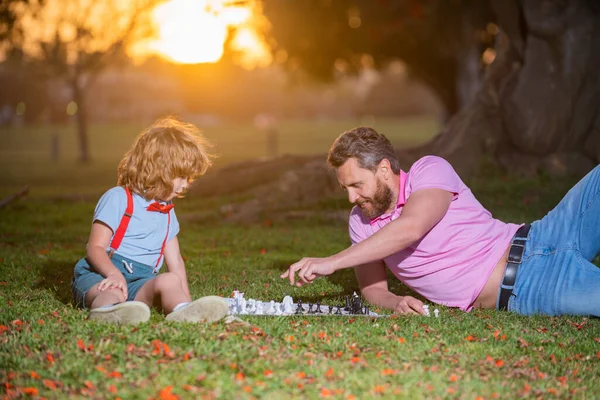 Father and son playing chess lying on grass at lawn park. Fathers Day, love family, parenthood, childhood concept. Active people having fun and playing chess at park, spend time with child. — Stock Photo, Image
