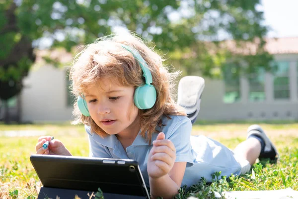 Liten skolpojke elev använder anteckningsbok eller surfplatta i parken på gräs. Självstudier, inlärning av barn och studier på skolgården. Tidig utveckling för barn. — Stockfoto