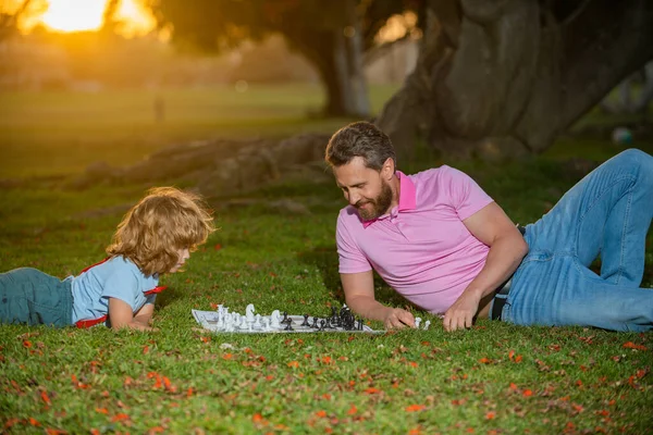 芝生の公園でチェスをしている父と息子。若い男の子がチェスで男を殴った。父の日、愛の家族、親、子供時代の概念. — ストック写真