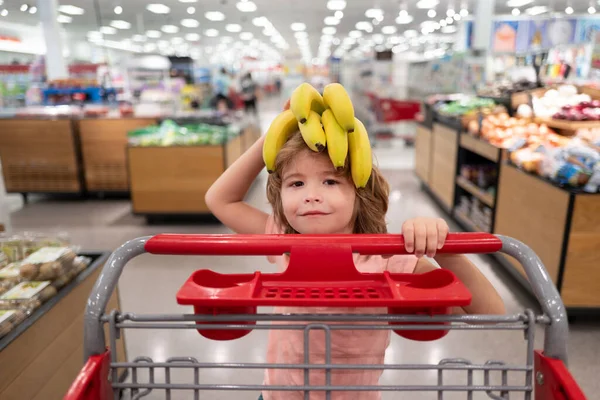 Забавный парень с тележкой, покупающий еду в продуктовом или супермаркете. Удивлённые дети. Банан на забавной детской голове. — стоковое фото