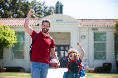 Öğretmenler günü. İlkokulun ebeveynleri ve öğrencileri el ele gider. Tişörtlü öğretmen ve okul parkının yanında sırt çantalı sevimli okul çocuğu..