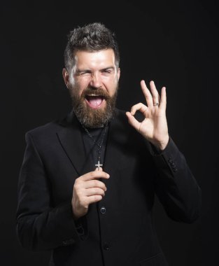 Beyaz arka plan üzerinde duran rahip üniforması giyen genç adam el hareketleriyle gülümsüyor ve mutlu yüzle parmaklarının arasından bakıyor..