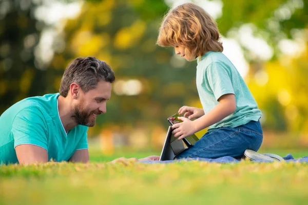 Ευτυχισμένος πατέρας που χρησιμοποιεί φορητό υπολογιστή χαλαρώστε με το γιο μαθητή κρατώντας φορητό υπολογιστή διασκεδάστε μαζί, χαμογελώντας ο μπαμπάς και το μικρό παιδί αγόρι απολαμβάνουν το Σαββατοκύριακο με gadgets έξω στη φύση. Βίντεο κλήση ενός εγγονού στους γονείς. — Φωτογραφία Αρχείου
