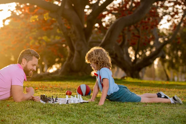 Father and son playing chess lying on grass at lawn park. Fathers Day, love family, parenthood, childhood concept. Child learning to play chess. Little boy think or plan chess game. Checkmate. — Stock Photo, Image