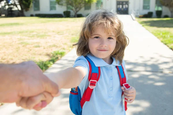Mały chłopiec z plecakiem trzymający ojców za rękę na rozmazanym szkolnym tle. Dziecko z plecakami stojące w parku koło szkoły. — Zdjęcie stockowe