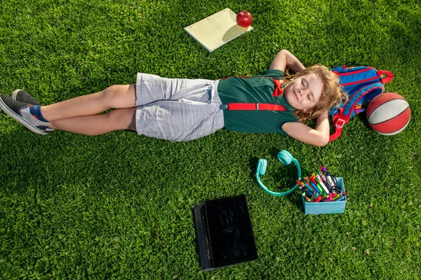 Heureux enfant profitant dans le parc pendant la journée d'été et d'automne dans la nature sur l'herbe verte avec fournitures scolaires en plein air. Vue du dessus du petit garçon étendu sur l'herbe verte. — Photo