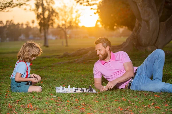 息子は草の上に寝そべって父親とチェスをする。男の子は親とチェスをする。認知発達。知的子供向けゲーム。賢い子供たち. — ストック写真