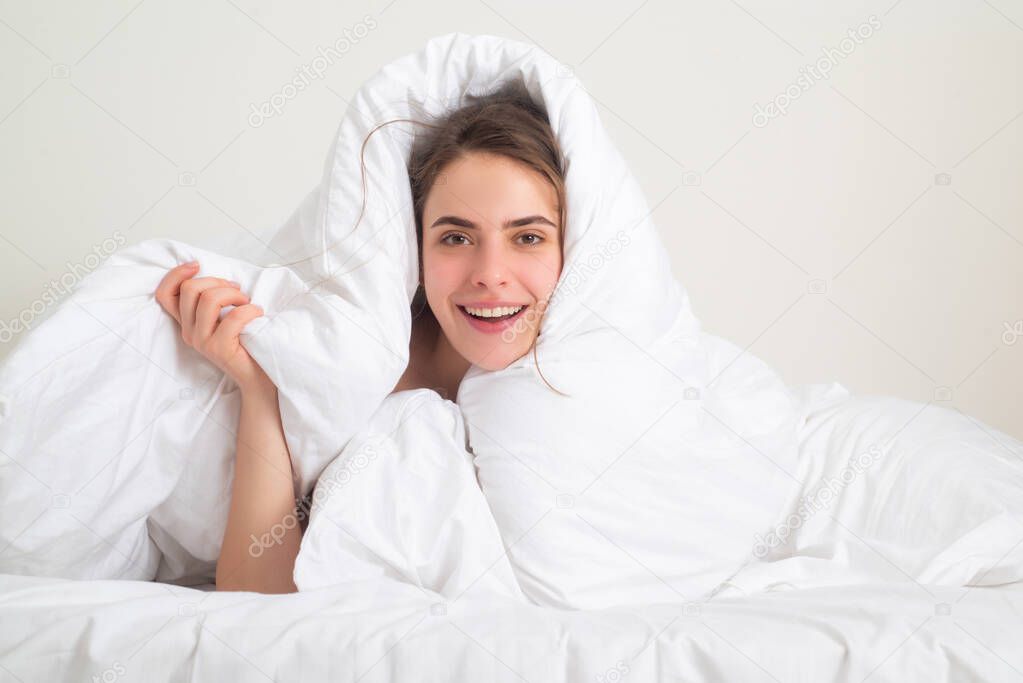 Funny woman with beautiful face is sitting on the bed, wrapped in soft blanket. Young woman lying in bed and hiding under the sheet.